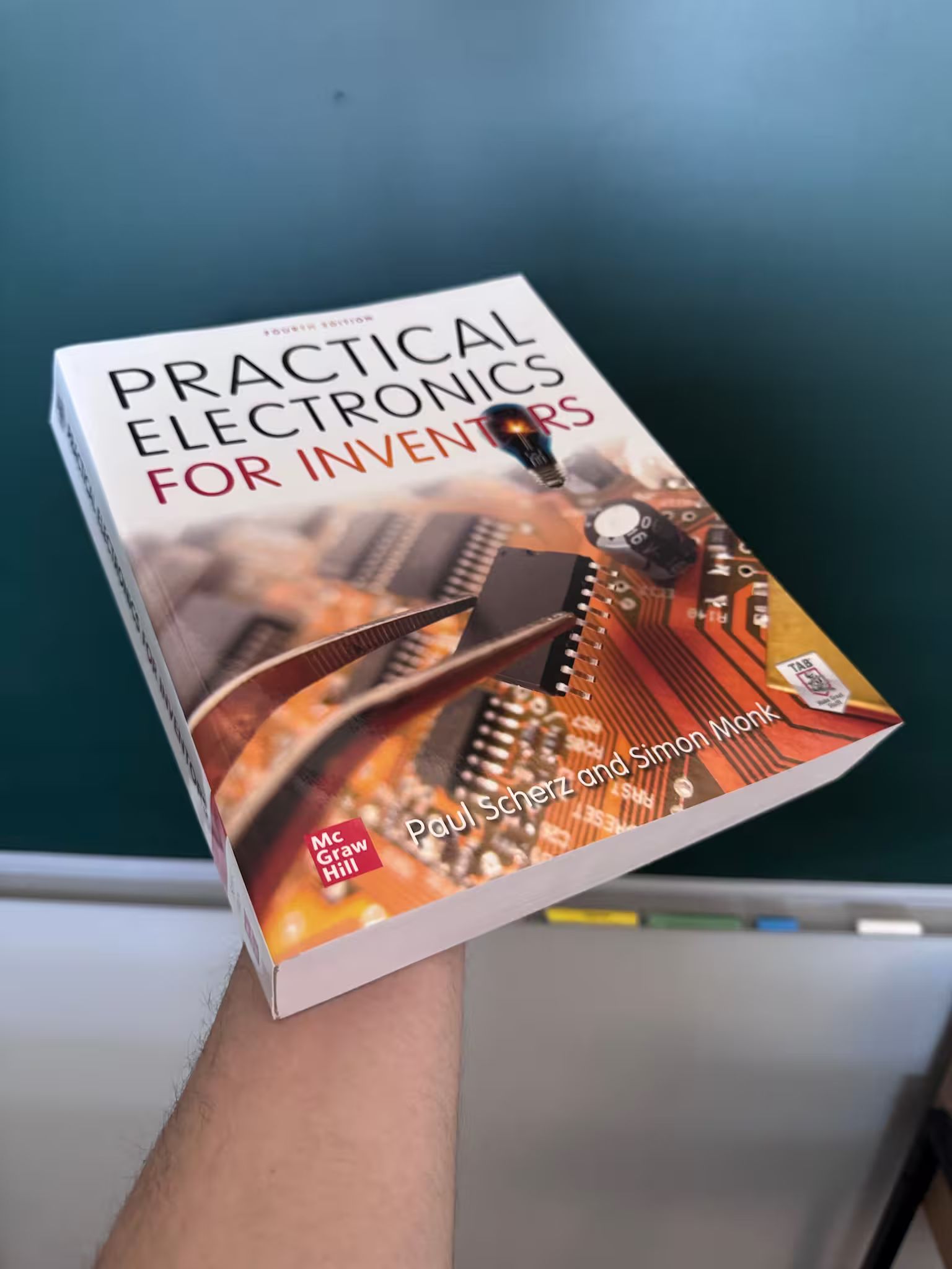 A hand holding up the thick fourth edition paperback of Practical Electronics for Inventors by Paul Scherz and Simon Monk.